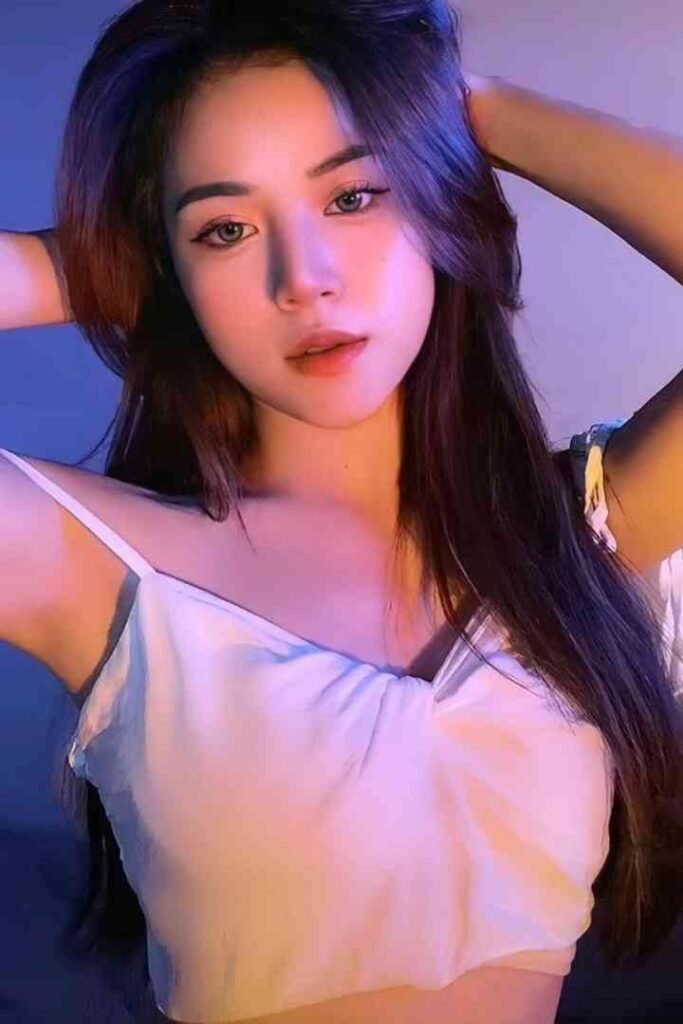 Young woman with long dark hair, wearing a white camisole, posing with hands behind head.