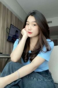 Young woman with long dark hair in a blue top and jeans, resting her chin on her hand.