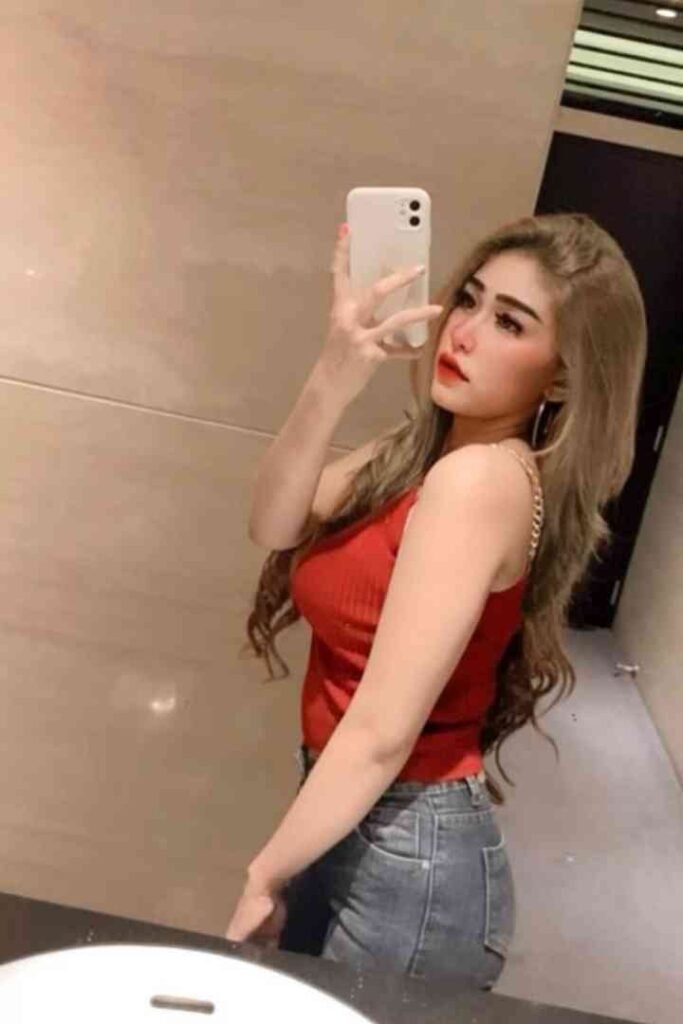 Woman with long blonde hair wearing red top and jeans taking a selfie in a bathroom mirror