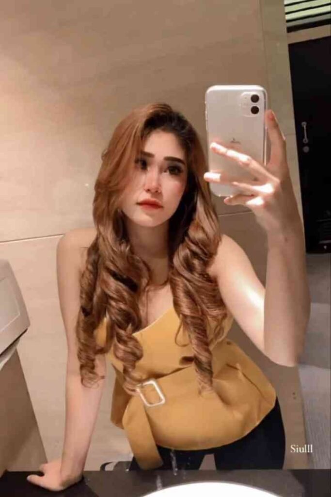 Young woman with long, wavy brown hair taking a mirror selfie in a yellow top.