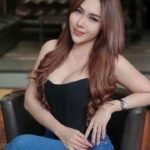 Woman with long brown hair wearing black corset and blue jeans sitting on black chair