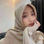 Young woman wearing a beige hijab and sweater, resting her chin on her hand.