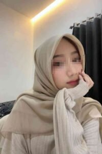 Young woman wearing a beige hijab and sweater, resting her chin on her hand.